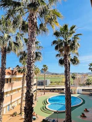 A pool surrounded by palm trees, a building with balconies, and a distant view of a mountain.