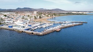Vista aérea de un puerto deportivo con barcos amarrados, una pequeña península con edificios y colinas al fondo, así como una ciudad distante.