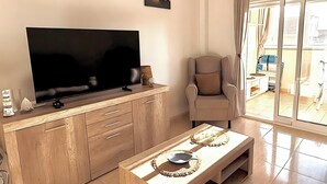 Sala de estar com televisão de tela plana, mesa de centro de madeira, poltrona bege e uma janela grande com cortinas.