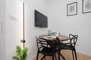 A modern room with a flat-screen TV mounted on the wall, a round wooden table with two black chairs, a green vase, and a framed botanical print. - A Functional Studio in the City Centre | Stary Browar (Poznań)