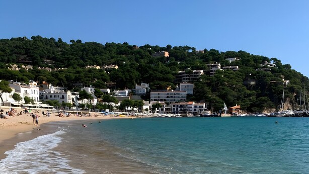 A beach with clear water, a sandy shore, and buildings in the background.