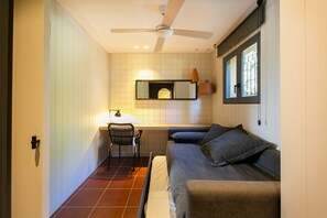 Une petite pièce comprenant un lit, une chaise, un bureau, un ventilateur de plafond, une fenêtre et un miroir.
