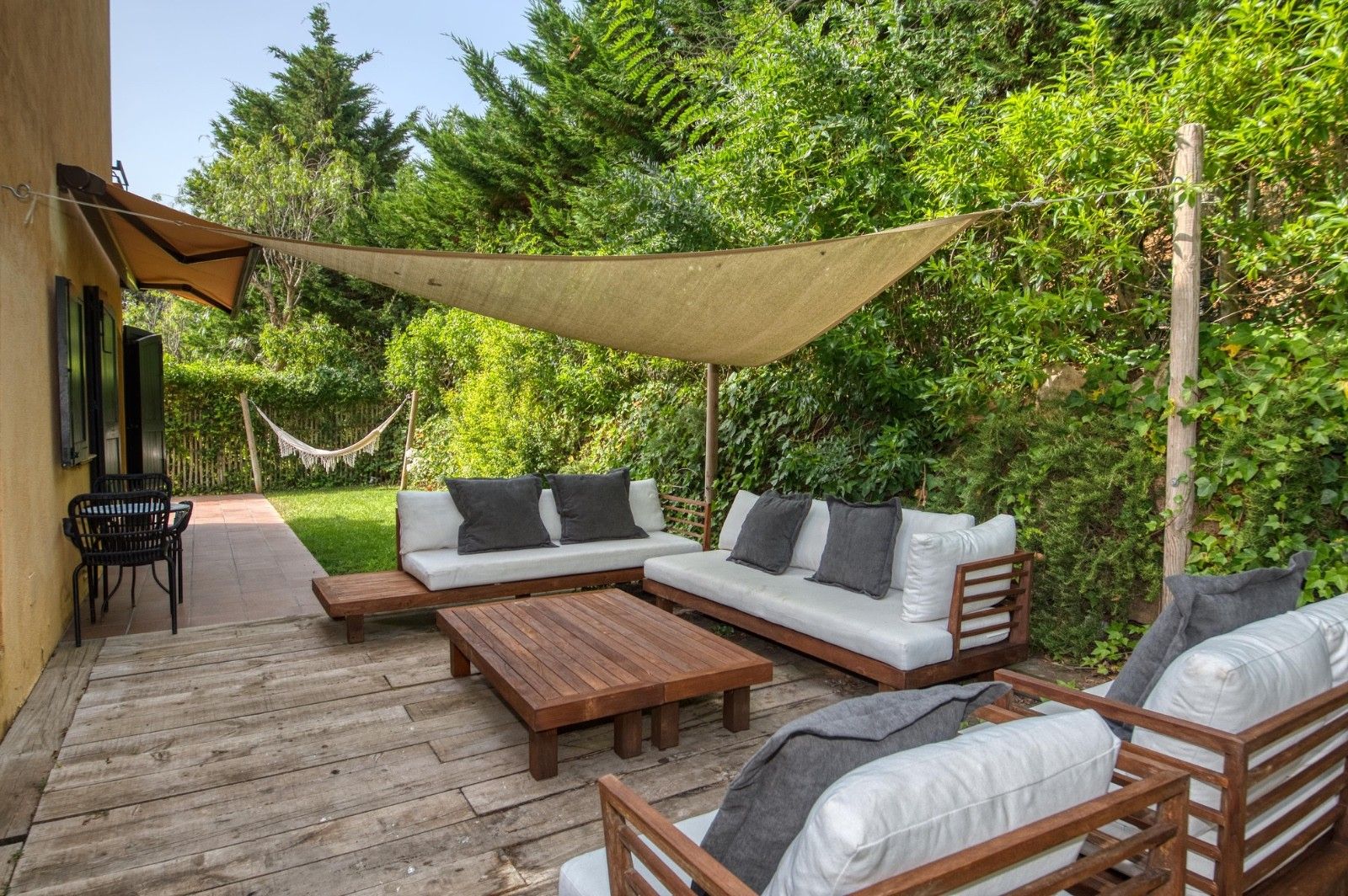 Un patio exterior con un conjunto de sofá, una mesa de madera y sillas, rodeado de follaje y una zona con sombra.