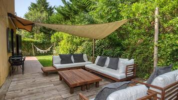Un patio exterior con un conjunto de sofá, una mesa de madera y sillas, rodeado de follaje y una zona con sombra.