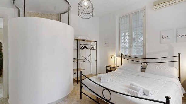 A bedroom with a bed, a round white column, a hanging light fixture, a window with blinds, and a wall-mounted air conditioner. - Apartment 'Portatrastevere' with Balcony, Wi-Fi and Air Conditioning (Rome)