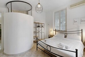 Une chambre à coucher avec un lit, une colonne blanche ronde, un luminaire suspendu, une fenêtre avec des stores et un climatiseur fixé au mur.