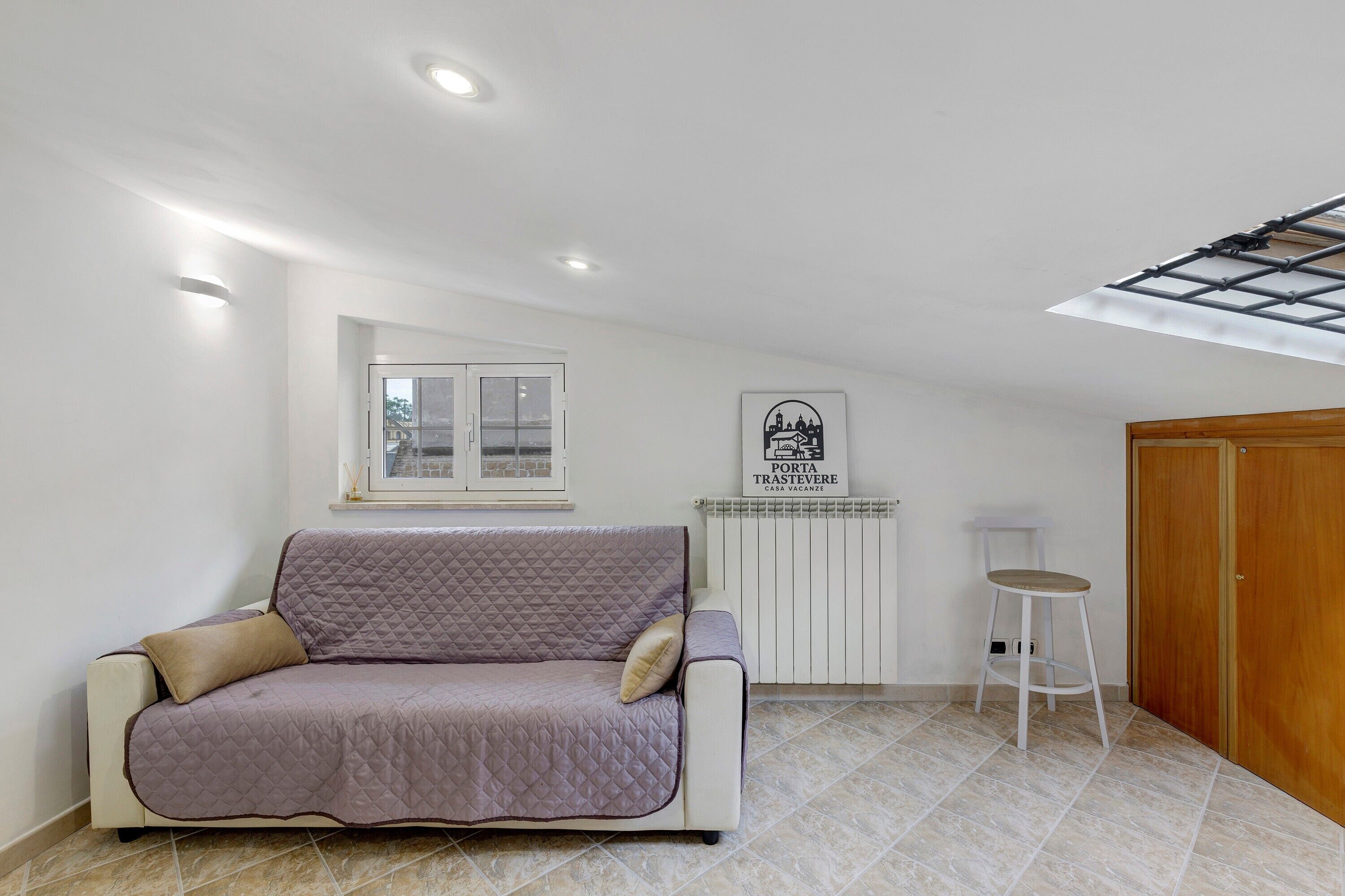Een kamer met een wit plafond, een raam, een witte radiator, een houten deur, een witte kruk en een paars gewatteerde bank.