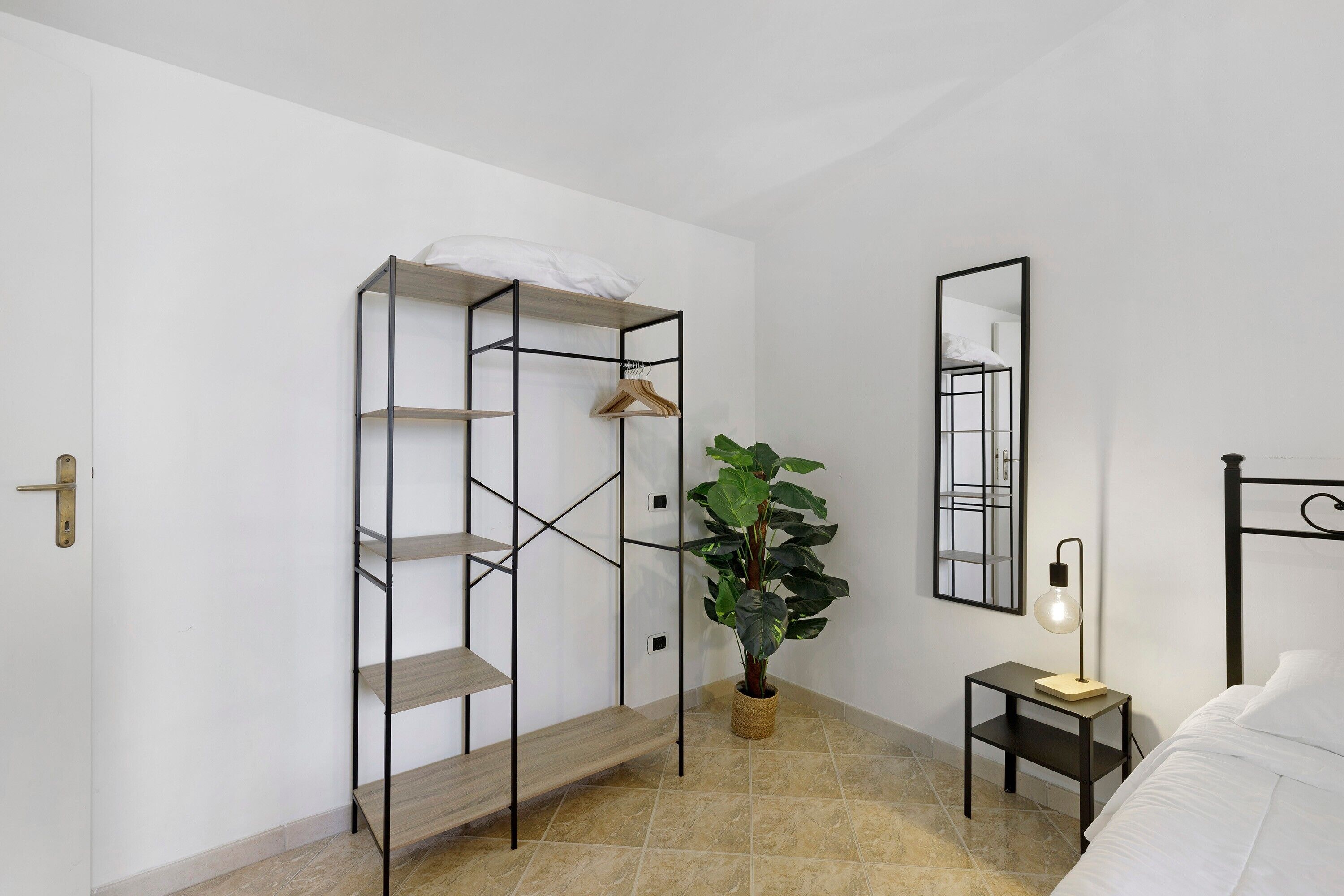 Een slaapkamer met een bed, een nachtkastje met een lamp, een spiegel aan de muur, een metalen schap met een hangende kledingstuk en een potplant.