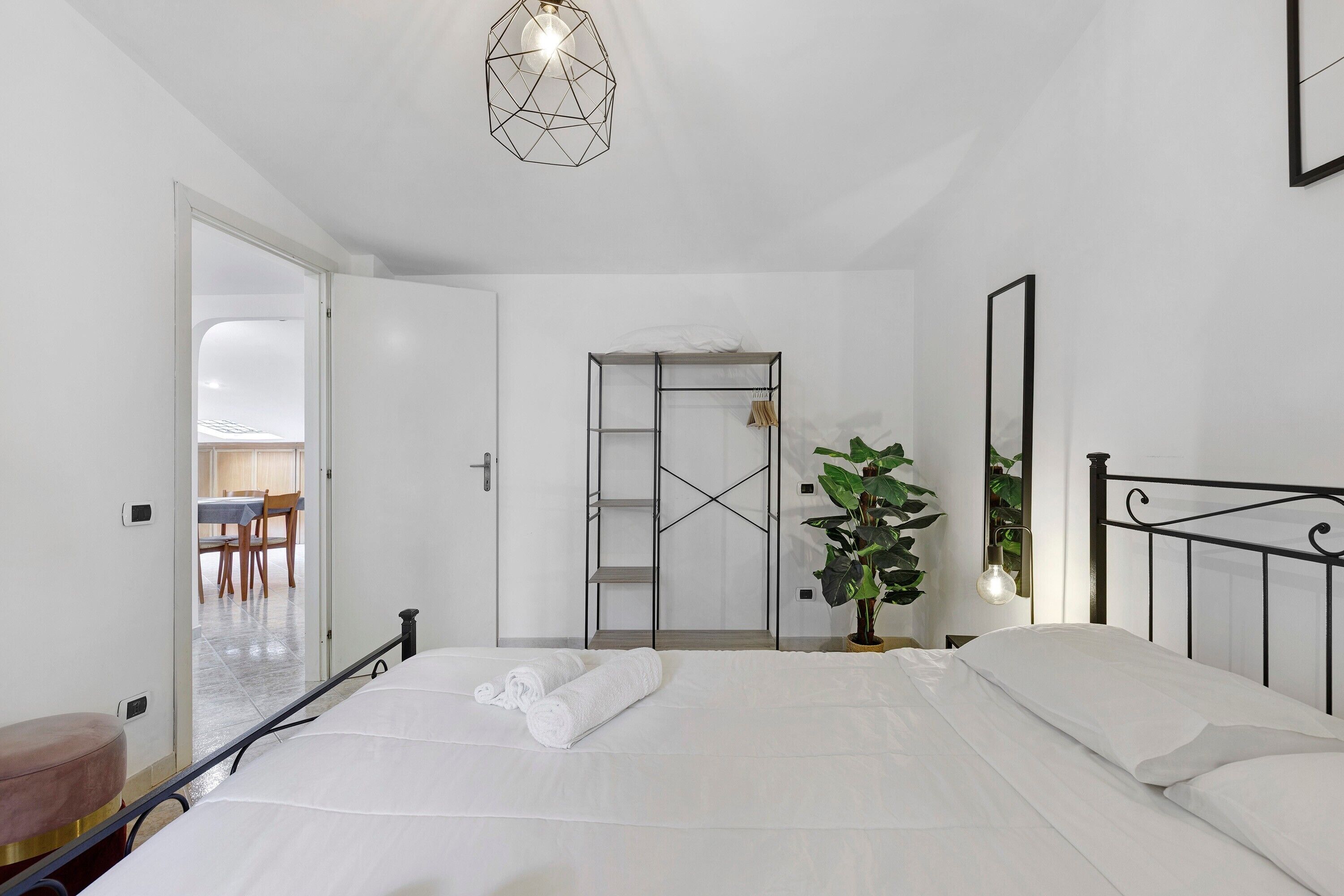 A bedroom with a bed, a door leading to a dining area, a potted plant, and a geometric light fixture.