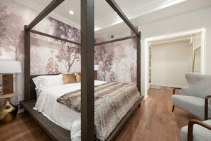 Uma cama com dossel com cobertor branco e marrom, uma poltrona cinza e uma parede com mural de árvore.