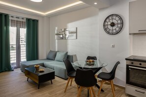 A modern living room with a glass dining table, black chairs, a sofa, a coffee table, a wall clock, and a kitchen area with an oven. - Apartment 'Ceramicist' with Shared Terrace, Wi-Fi and Air Conditioning (Heraklion)