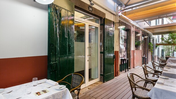 Uma área de refeições ao ar livre com mesas e cadeiras, entrada por uma porta de vidro e um deck de madeira.