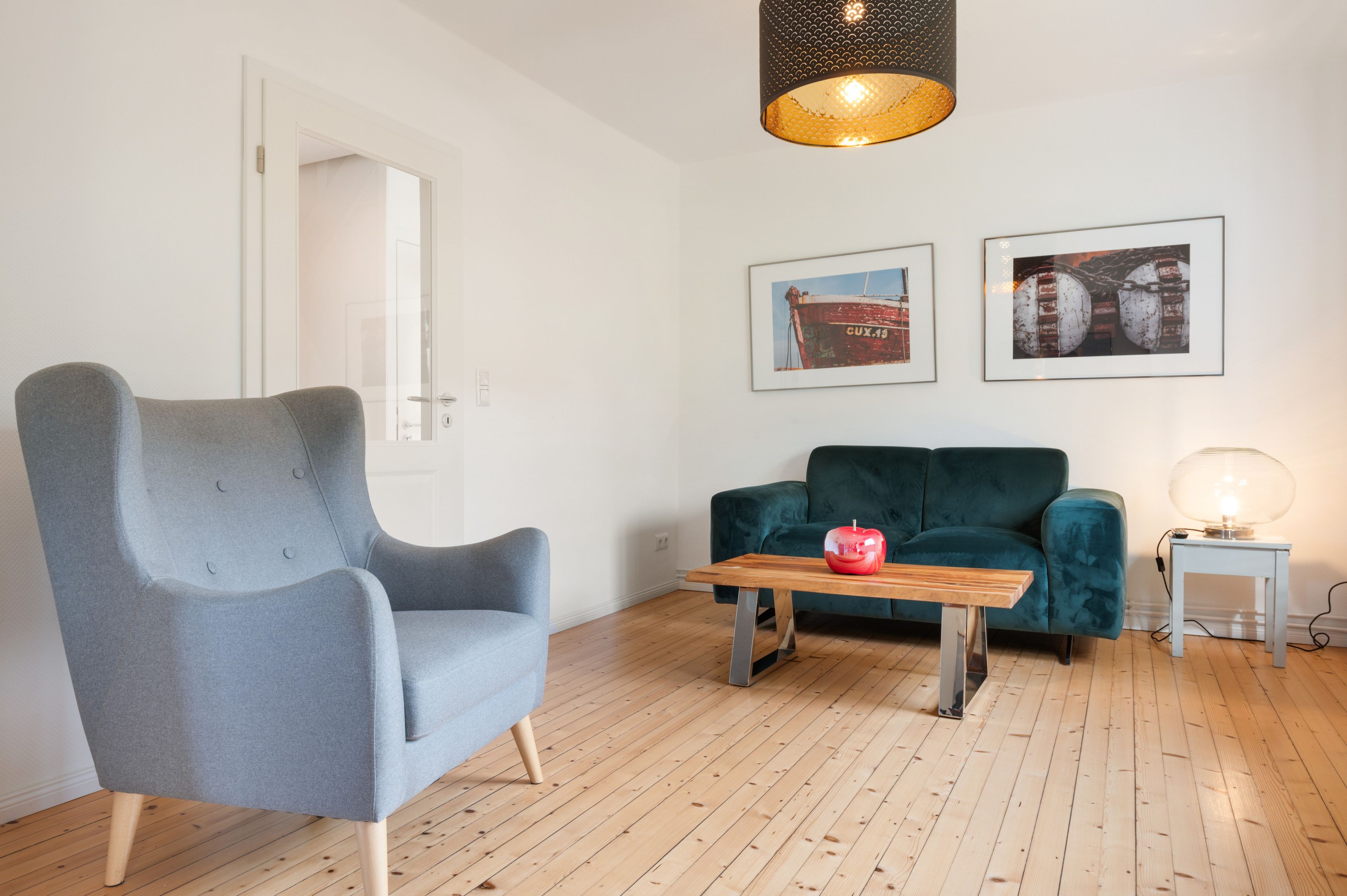 A room with a grey armchair, a teal sofa, a wooden coffee table with a red apple, and a hanging light fixture.
