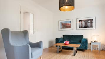 A room with a grey armchair, a teal sofa, a wooden coffee table with a red apple, and a hanging light fixture.
