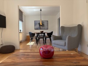Ein modernes Wohnzimmer mit Holzboden, einer roten, kugelförmigen Skulptur auf einem Couchtisch, einem grauen Sessel, einem Esstisch mit Stühlen und einem Gemälde an der Wand.