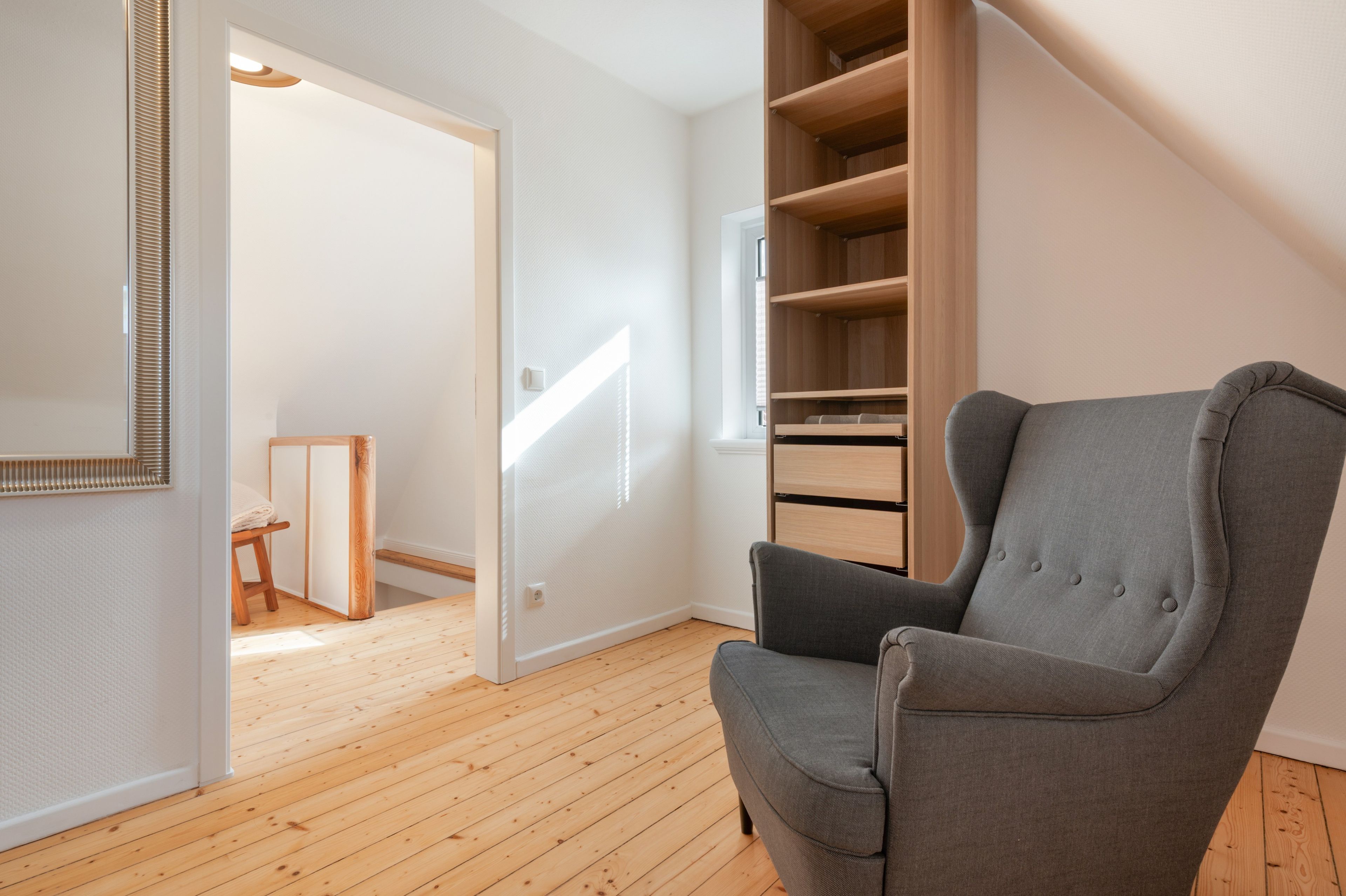 Une pièce avec un fauteuil gris, un plancher en bois, une porte blanche et une bibliothèque intégrée en bois.
