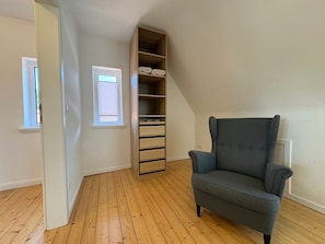 Ein Zimmer mit Holzboden, einem grauen Sessel, einer eingebauten Holzregalanlage und einem Fenster mit Jalousien.