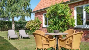 Een terras met rieten meubels en een tafel, omringd door een stenen huis en begroeiing.