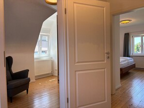 Ein Zimmer mit Holzboden, einem schwarzen Stuhl, einer weißen Tür und einem Bett mit Blick aus dem Fenster.
