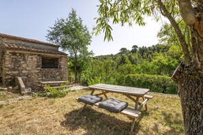 Een rustiek stenen gebouw met een houten tafel en bankjes in een grasrijke omgeving.