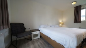 Ein Schlafzimmer mit einem Bett, einem Sessel, einem Nachttisch, einer Lampe und einem Fenster mit Vorhängen.