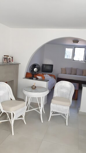Um espaço compacto para viver com cama, televisão, ventoinha e duas cadeiras de vime em torno de uma mesinha.