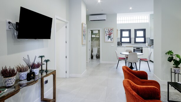 A modern living room with a flat-screen TV, a glass coffee table with decorative items, and a seating area with orange chairs.