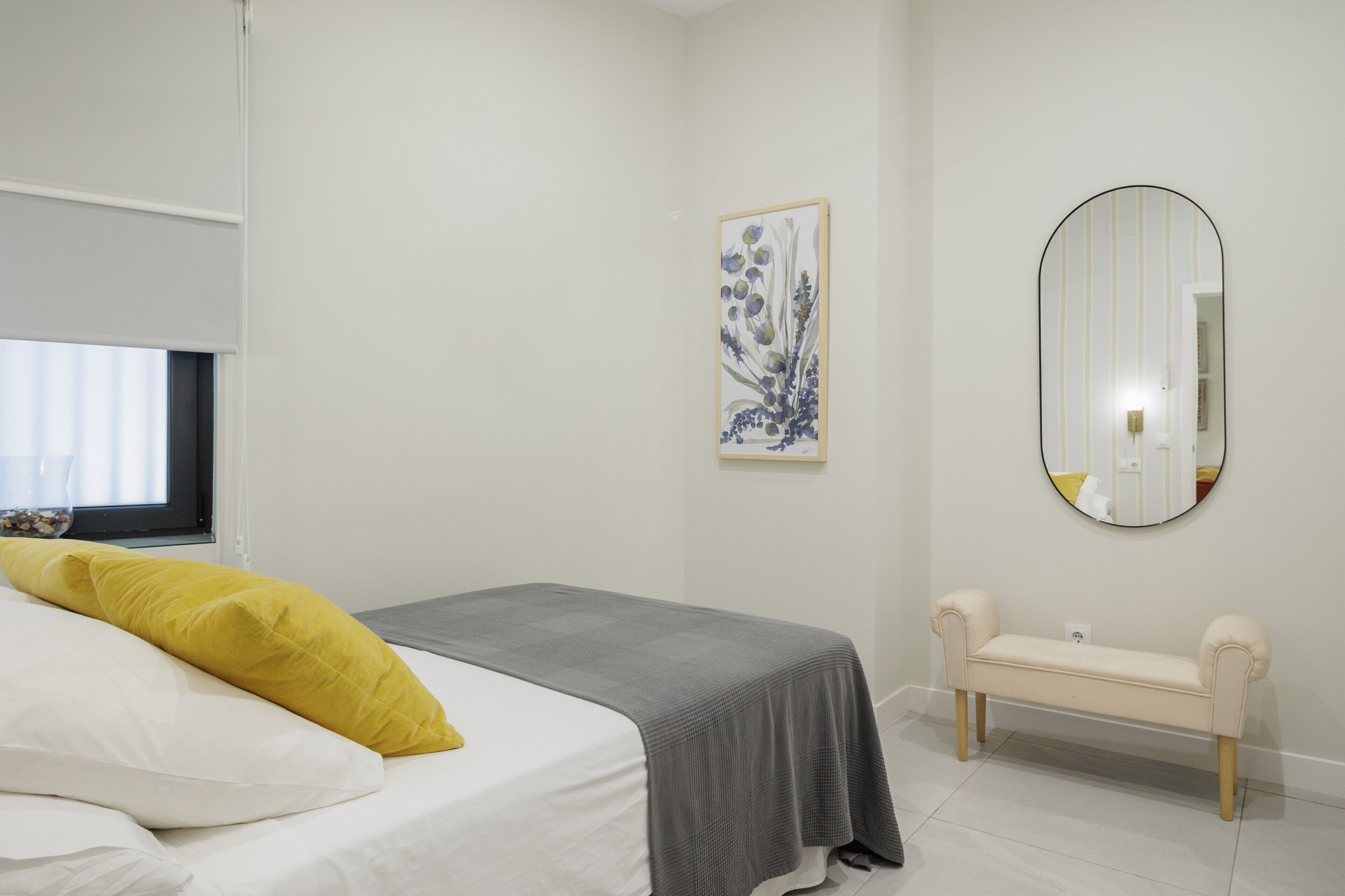 A bedroom with a bed, a bench, a wall-mounted mirror, and a framed floral artwork.