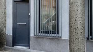 A modern building facade with a dark-colored door, a window with vertical bars, and textured columns.