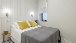 A neatly made bed with white and yellow pillows, a grey blanket, and a small side table with a basket.