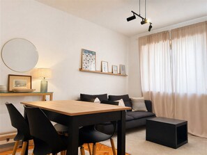 Ein modernes Wohnzimmer mit einem Holzesstisch, schwarzen Stühlen, einem grauen Sofa, einem schwarzen Hocker und einem Deckenprojektor.