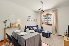 Ein modernes Wohnzimmer mit Esstisch für vier Personen, einer Couch, einem Fernseher und einem Fenster mit Vorhängen.