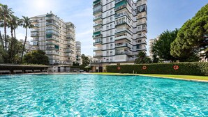 A swimming pool with clear water, surrounded by multi-story residential buildings, palm trees, and a hedge.