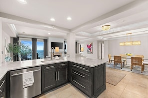 A modern kitchen with dark cabinetry, a large island, and a dining area with wicker chairs. - Relax Above the Emerald Coast | Luxury 2BR Gulf-Front Retreat (Panama City Beach)