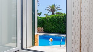 A window view of a swimming pool with a palm tree and hedge in the background. - Holiday Home 'Villa Jaume' with Private Pool, Private Garden and Air Conditioning (Cap d'Artrutx)