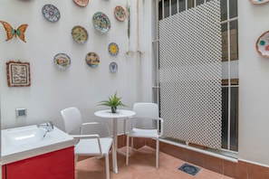 Un petit espace patio avec un évier blanc, deux chaises blanches, une petite table avec une plante et des assiettes décoratives accrochées au mur.