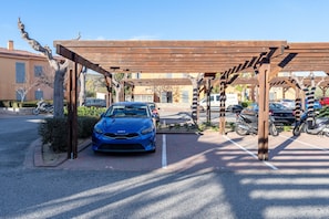 Een parkeerplaats met een blauwe auto onder een houten pergola, omringd door geparkeerde motorfietsen en auto's.