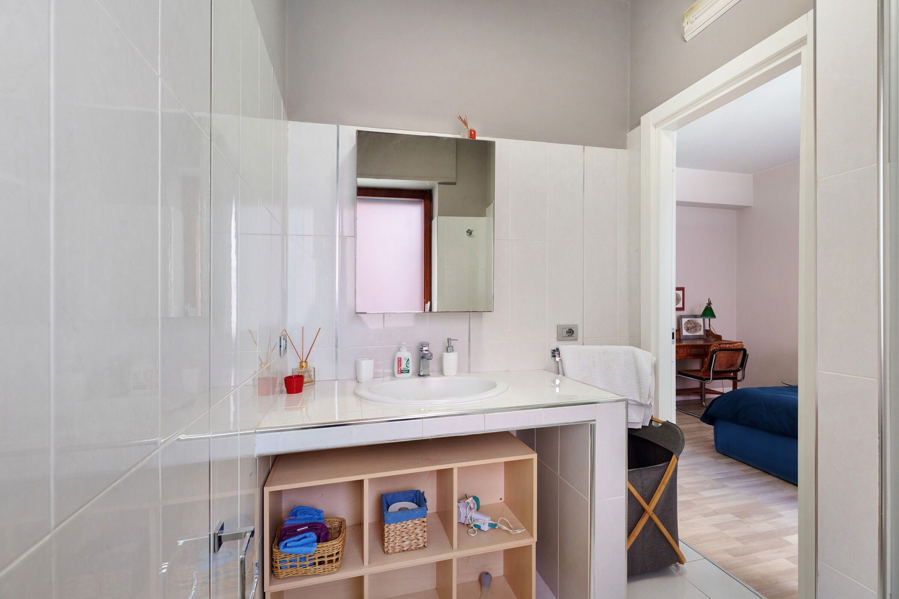 A modern bathroom with a glass vanity, a mirror, and a sink. There is a shelf with baskets and a towel rack.