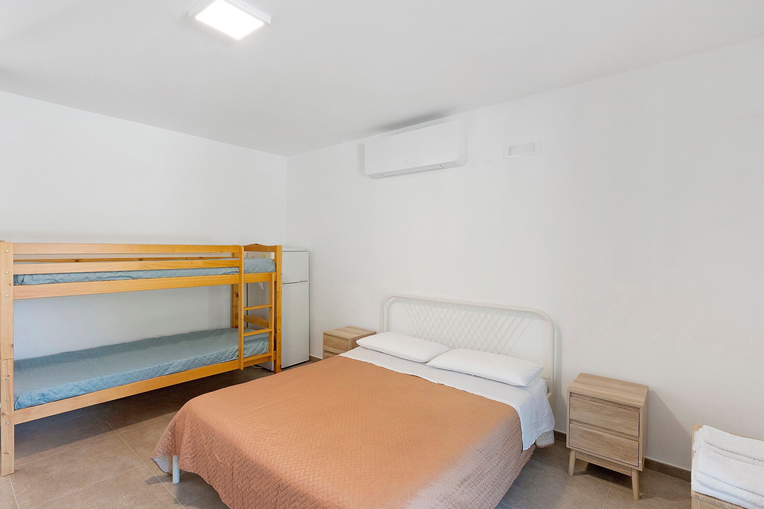 Een kamer met een stapelbed, een enkel bed, een nachtkastje en een wandairconditioner.