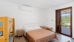 A bedroom with a bunk bed, a single bed, a wooden nightstand, a small table, and a sliding glass door leading to an outdoor area.