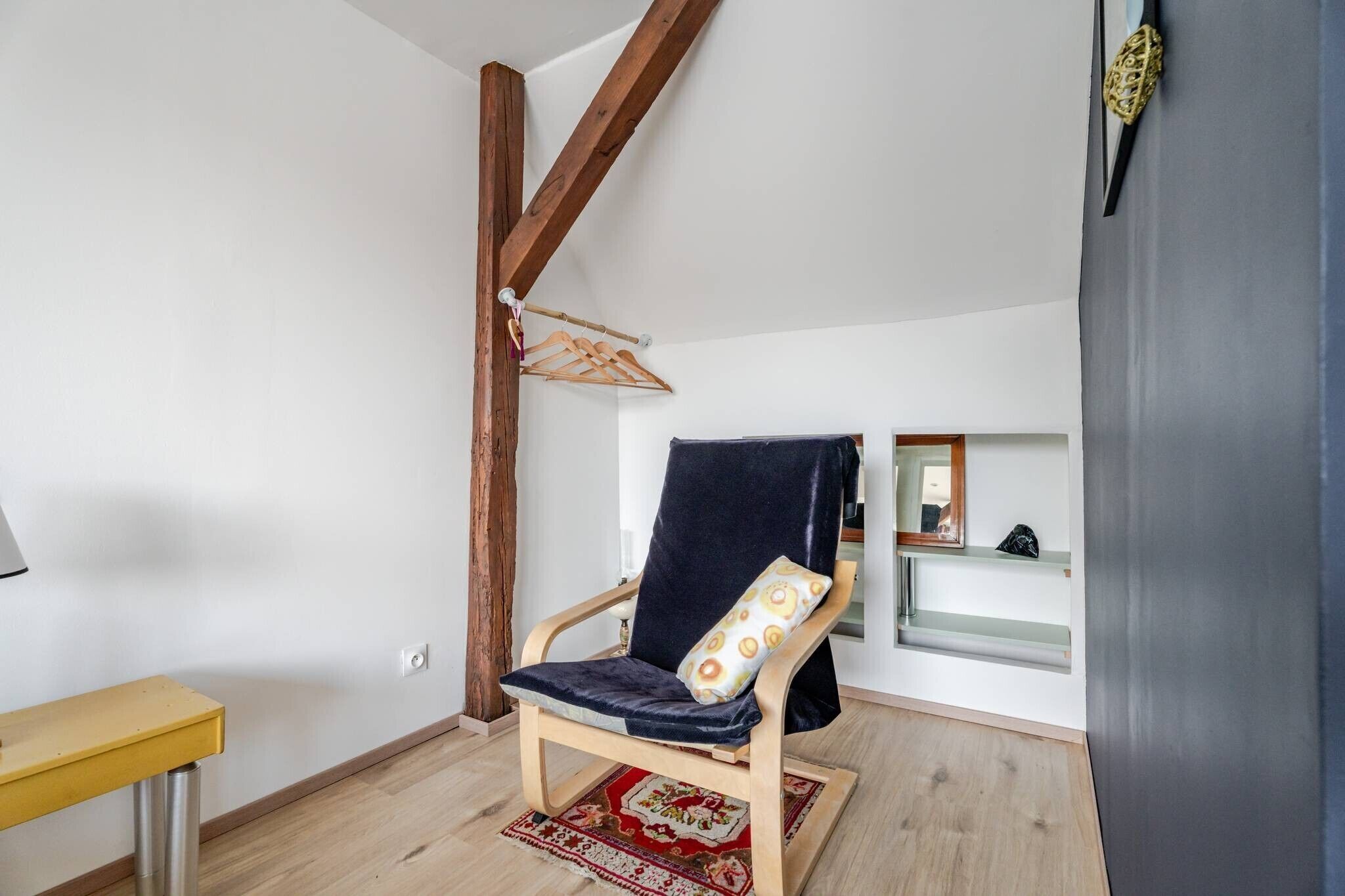 A room with a wooden chair, a small table, a hanging rack, and a mirror.