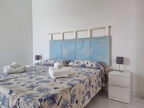 Una cama doble con una colcha azul y blanca, dos toallas blancas, una mesita de noche con una lámpara y un mueble auxiliar blanco.