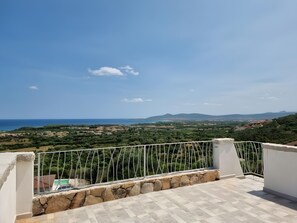 Une terrasse dotée d’un mur en pierre et d’une balustrade en métal, offrant une vue sur un paysage côtier verdoyant et sur des montagnes lointaines.