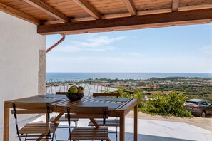 A wooden table with chairs and a bowl of fruit, overlooking a scenic landscape with a view of the sea and hills.