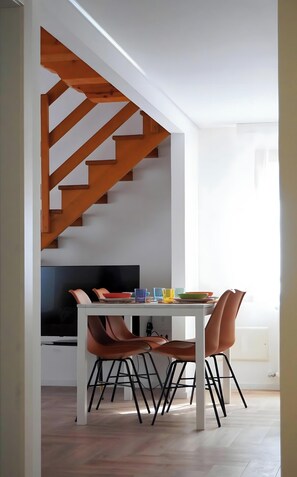 Un espace repas moderne avec une table blanche, des chaises marron, un téléviseur et un escalier en bois.