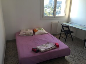 Een eenpersoonsbed met een roze sprei, twee kussens, een bureau met een stoel en een raam met uitzicht op gebouwen.