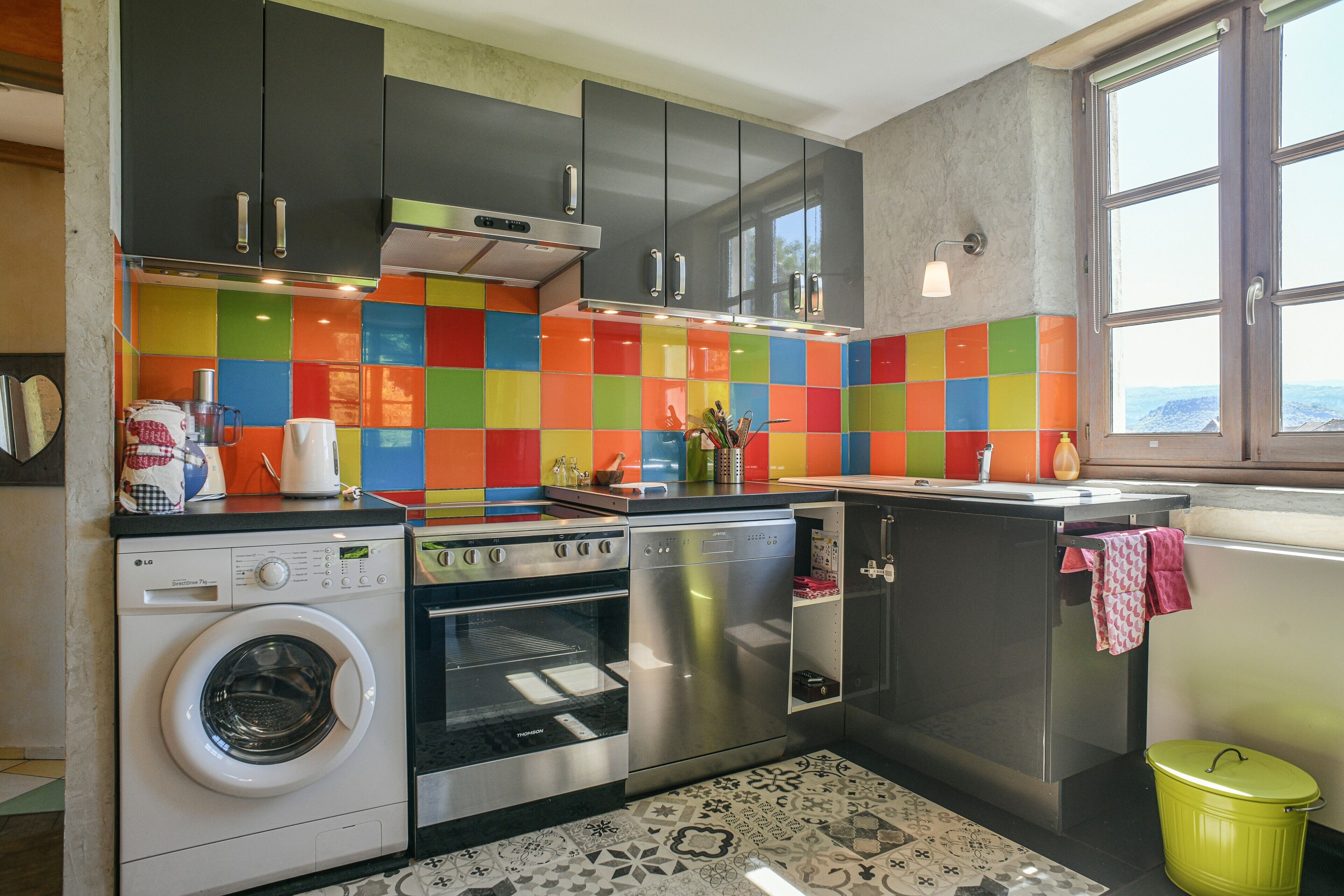 Une cuisine compacte équipée d’un lave-linge, d’un four et d’un plan de travail coloré recouvert de carreaux.