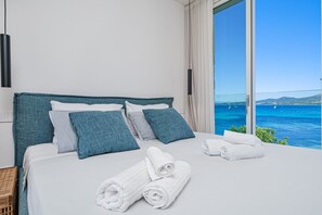 Ein Hotelzimmer mit einem großen Bett, weißen Leinentüchern, türkisfarbenen Kissen und Meerblick.