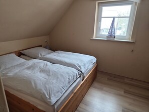 Ein Zimmer mit schrägem Dach, einem Bett mit Holzbettgestell, einem kleinen Fenster mit Blick auf Bäume und einem an der Wand befestigten Regal mit einer Lampe.