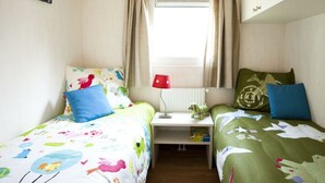 Ein Zimmer mit zwei Betten, einem fensterbelaubten Fenster, einem Heizkörper und einem kleinen Tisch mit Lampe.